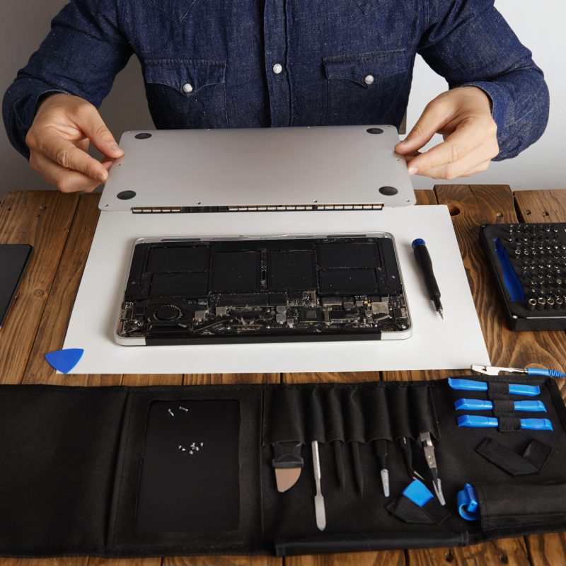 Service man opens backside topcase cover of computer laptop before repairing, cleaning and fixing it with his professional tools from toolkit box near on wooden table front view